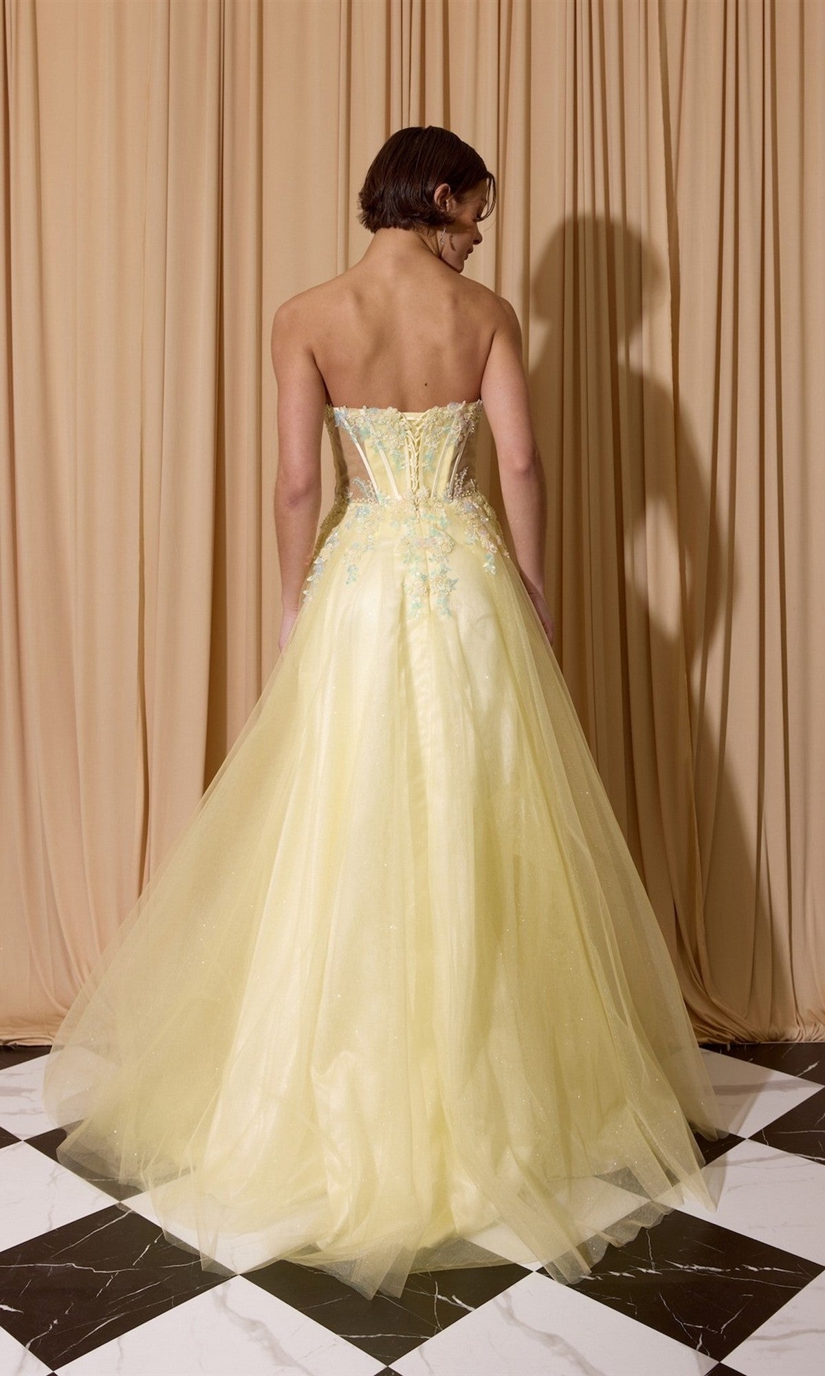 Standing on a black and white tiled floor, a person faces away from the camera against a beige curtain backdrop, wearing the Eureka EK208 Long Prom Dress—a light yellow strapless gown with a fitted bodice and full glitter tulle skirt.